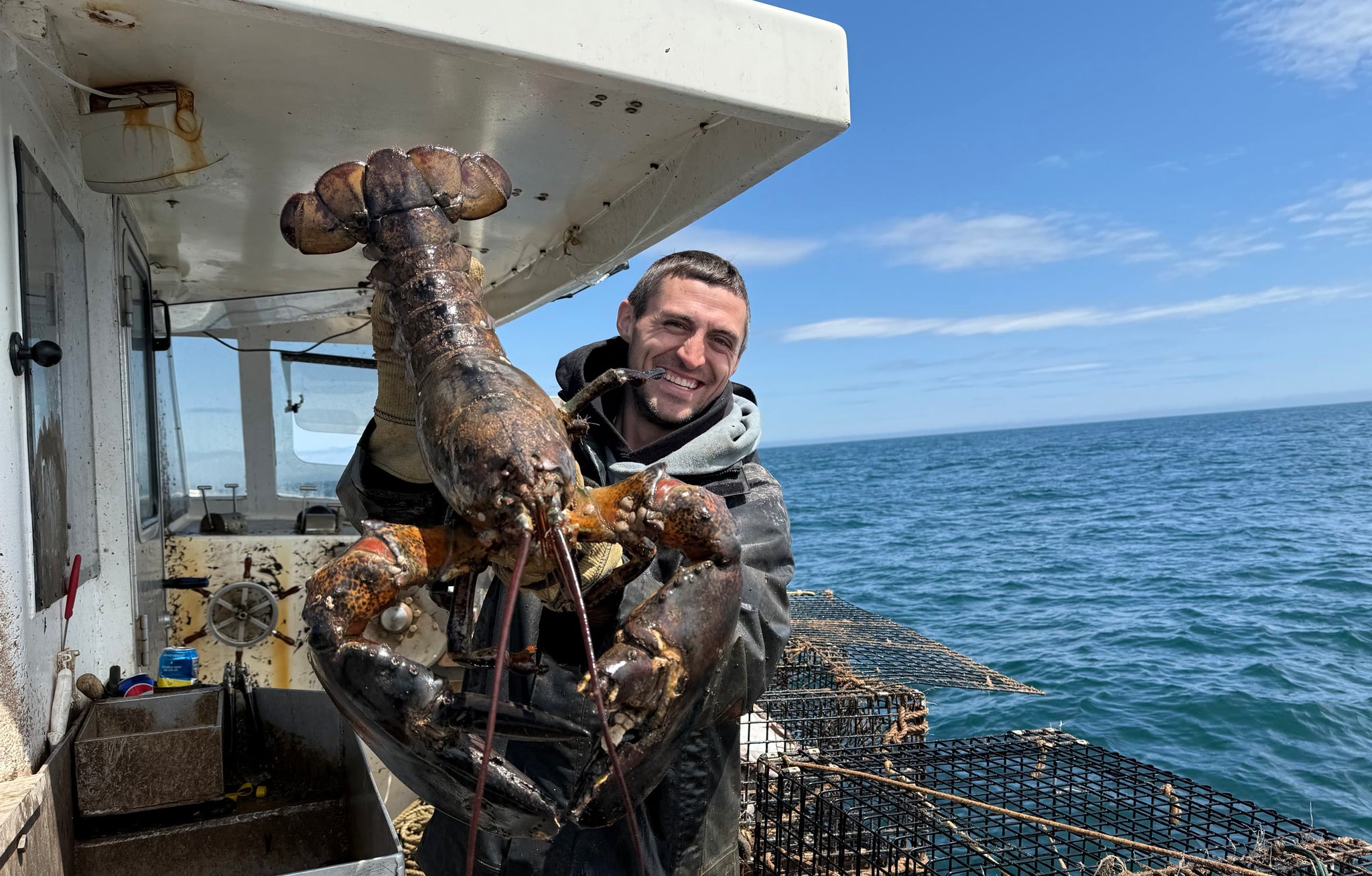 Jacob holding a barnacled lobster on deck under a clear sky, traps stacked behind him